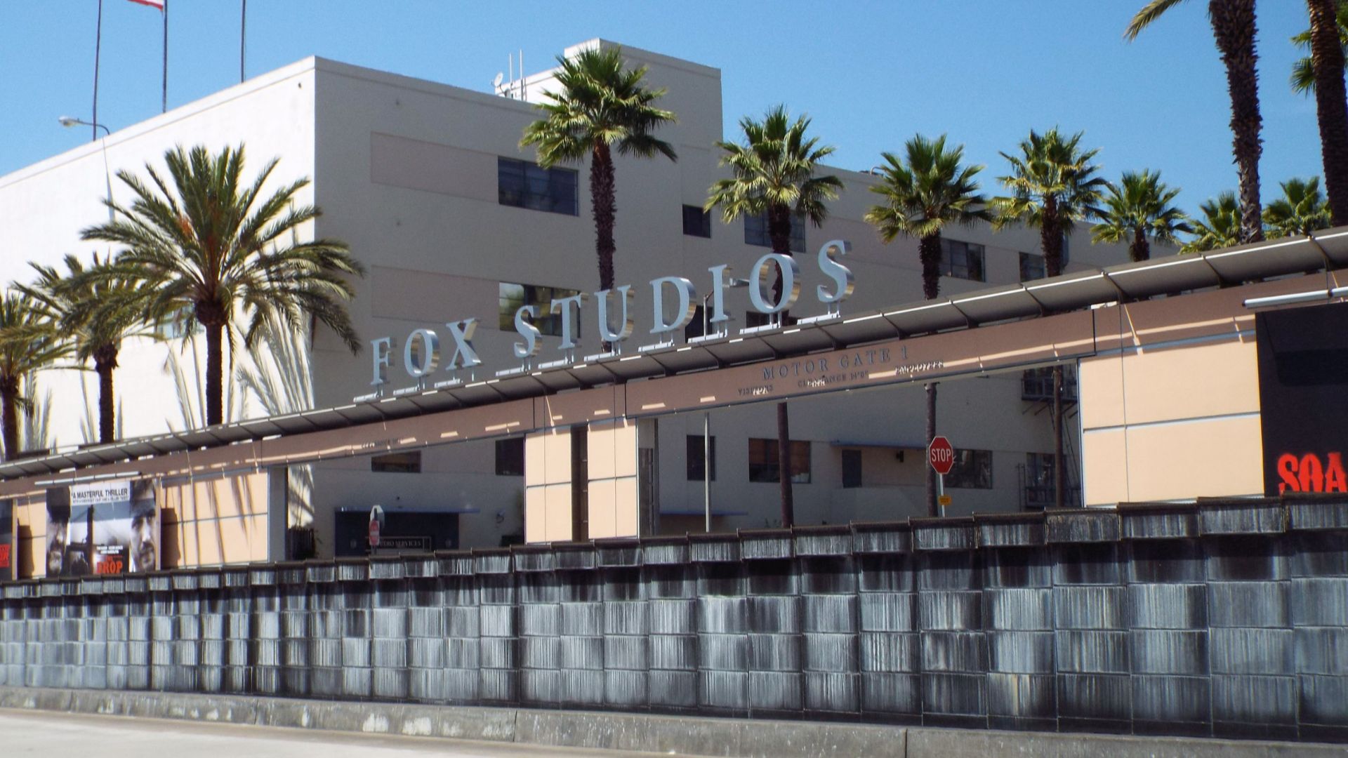 Entrance to the studio lot of 20th Century Fox in Century City, California as seen by passing traffic on Pico Boulevard. The black wall in front of the entrance gate is a retaining wall. Traffic entering or leaving the studio goes through a very sharp S-c