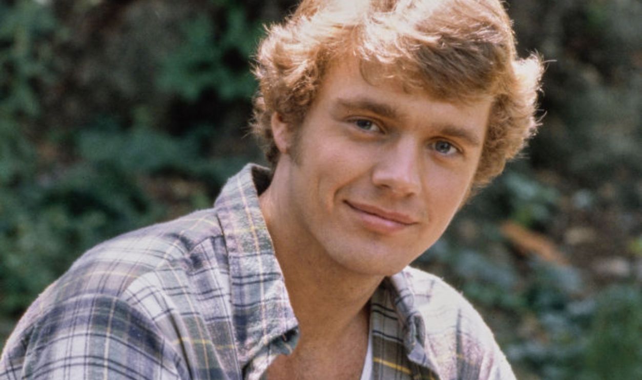 American actor and singer John Schneider, wearing an unbuttoned plaid shirt over a white t-shirt with jeans, United States, circa 1980.