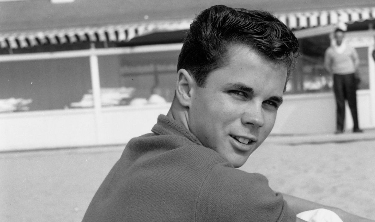 American actor Tony Dow looks over his shoulder while sitting on a beach, Santa Monica, California. Dow wears a casual top and light-colored pants rolled up at the cuffs.