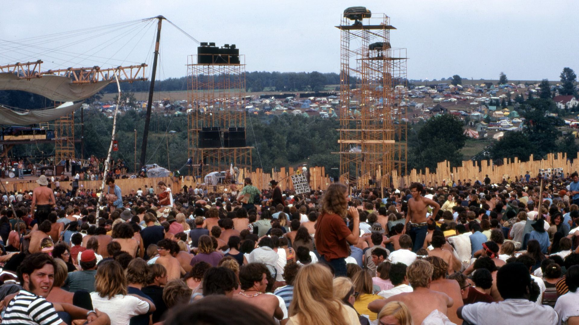 Picture taken on 17 August 1969 at the Woodstock Music and Art Fair. The person carrying the placard is Moonfire Lewis Beach Marvin III. Joe Cocker is performing on stage.