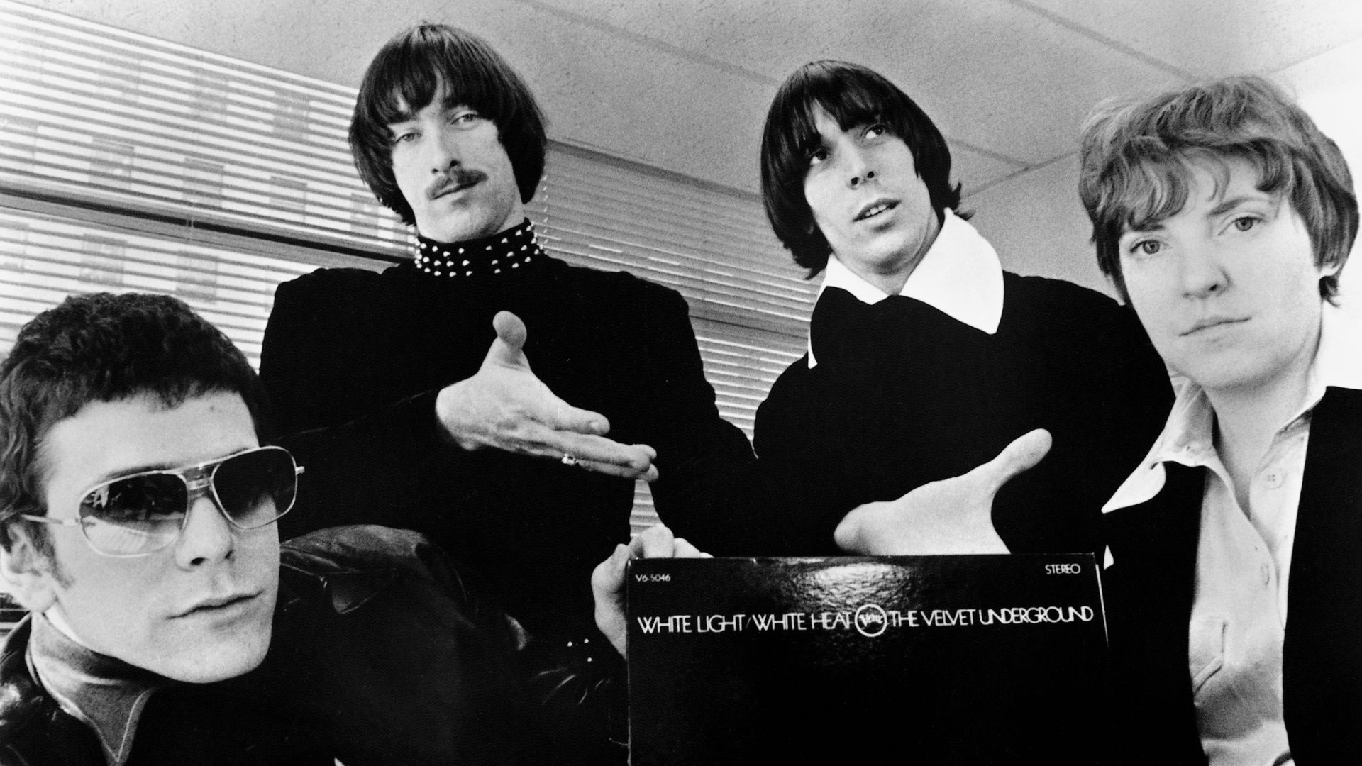 A publicity photo of the American rock band The Velvet Underground circa 1968, promoting their second album White Light/White Heat. The band members are positioned around a copy of the album, with the text of the title visible against a black background. From left to right: Lou Reed, Sterling Morrison, John Cale, and Maureen Tucker.