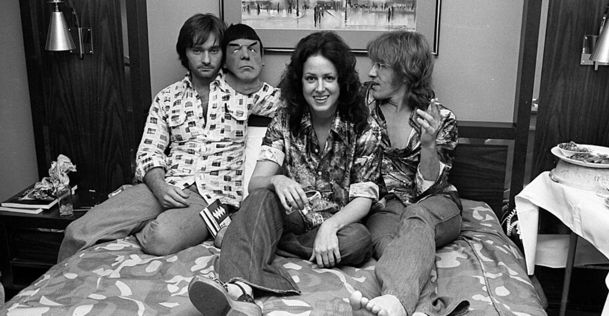 Marty Balin, Grace Slick, and Paul Kantner of Jefferson Starship seated on hotel room bed with a Mr. Spock mask in Los Angeles, California