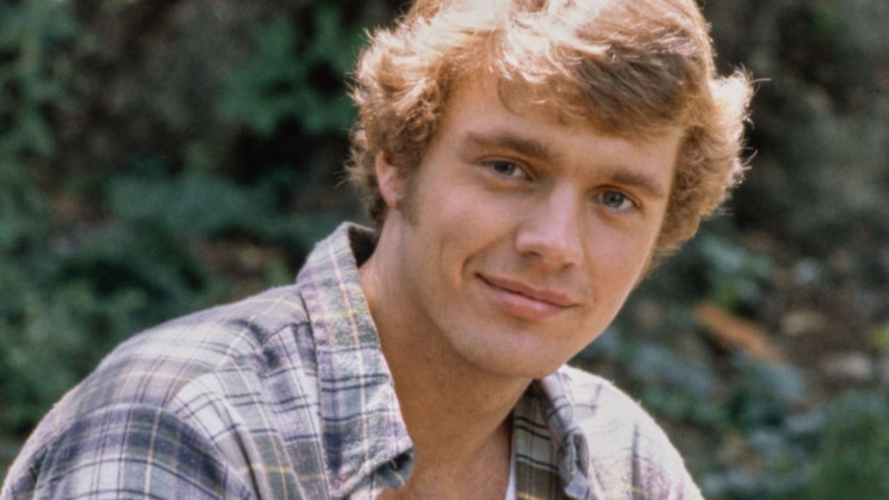 American actor and singer John Schneider, wearing an unbuttoned plaid shirt over a white t-shirt with jeans, United States, circa 1980.