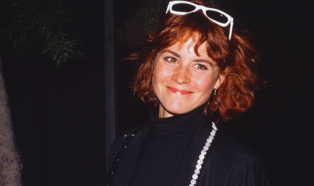 Ally Sheedy circa 1989: American actor Ally Sheedy smiles while wearing a long black coat and white-framed sunglasses on top of her head.
