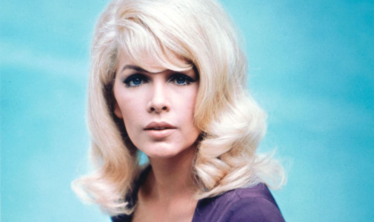 Stella Stevens, US actress, wearing a low-cut dark blue top in a studio portrait, against a light blue background