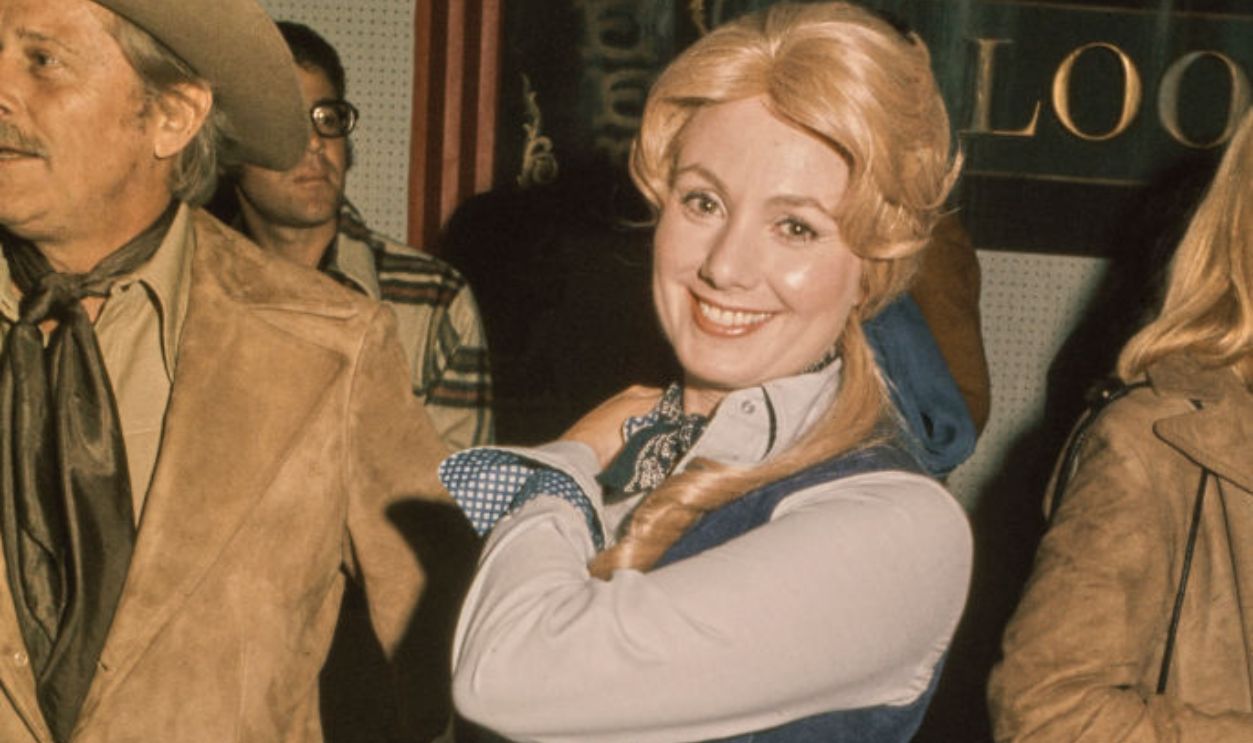American actress and singer Shirley Jones wearing a blue denim waistcoat over a white shirt with blue trim on the cuffs, and a blue denim skirt, adjusting a shoulder strap over her right shoulder, with a man in a suede jacket and a cowboy hat in the background, at an event, United States, circa 1973.