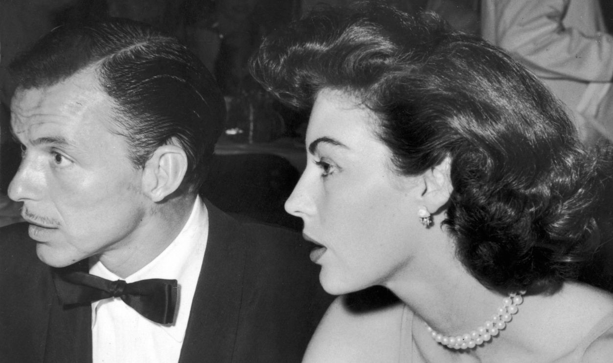 Getty Images - 3202821 - 1952: Profile view of singer Frank Sinatra, with a pencil-thin mustache, and his wife, actor Ava Gardner, facing an unseen person(s) while seated together, at the Flamingo resort. Sinatra wears a tuxedo; Gardner wears an evening dress with pearls.
