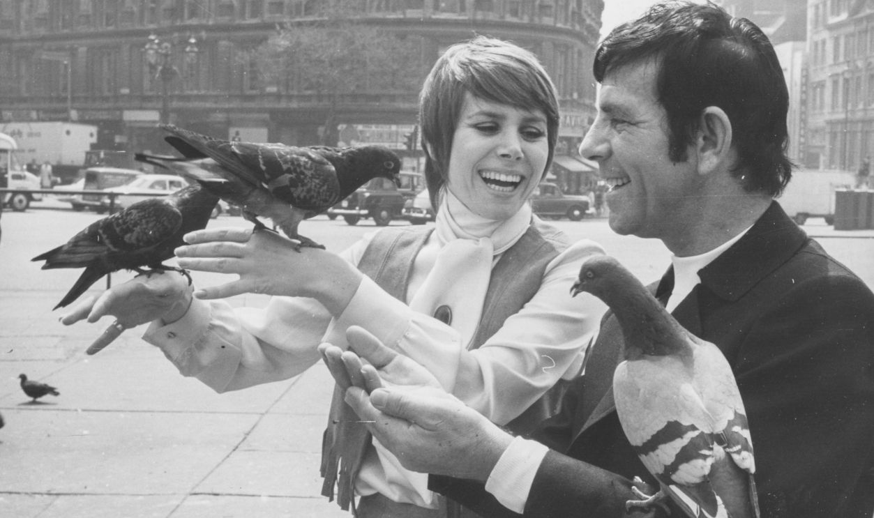 Actress Judy Carne and comedian Norman Wisdom feeding pigeons in Trafalgar Square as they promote their film 'All The Right Noises', London, May 12th 1969.