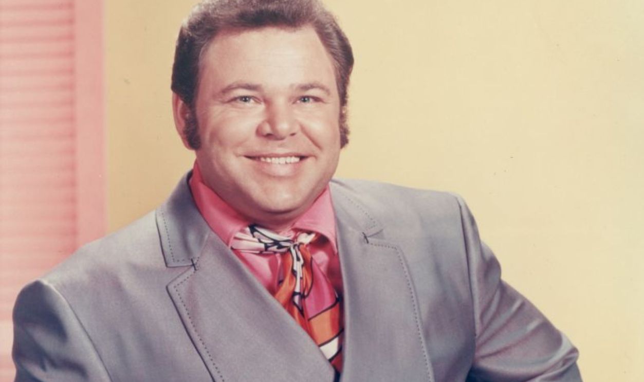 circa 1975: American country singer and television host Roy Clark poses with his hand on his hip in a promotional portrait for the CBS-TV series 'Hee Haw'. He wears an iridescent gray suit with a pink shirt and printed silk tie. 