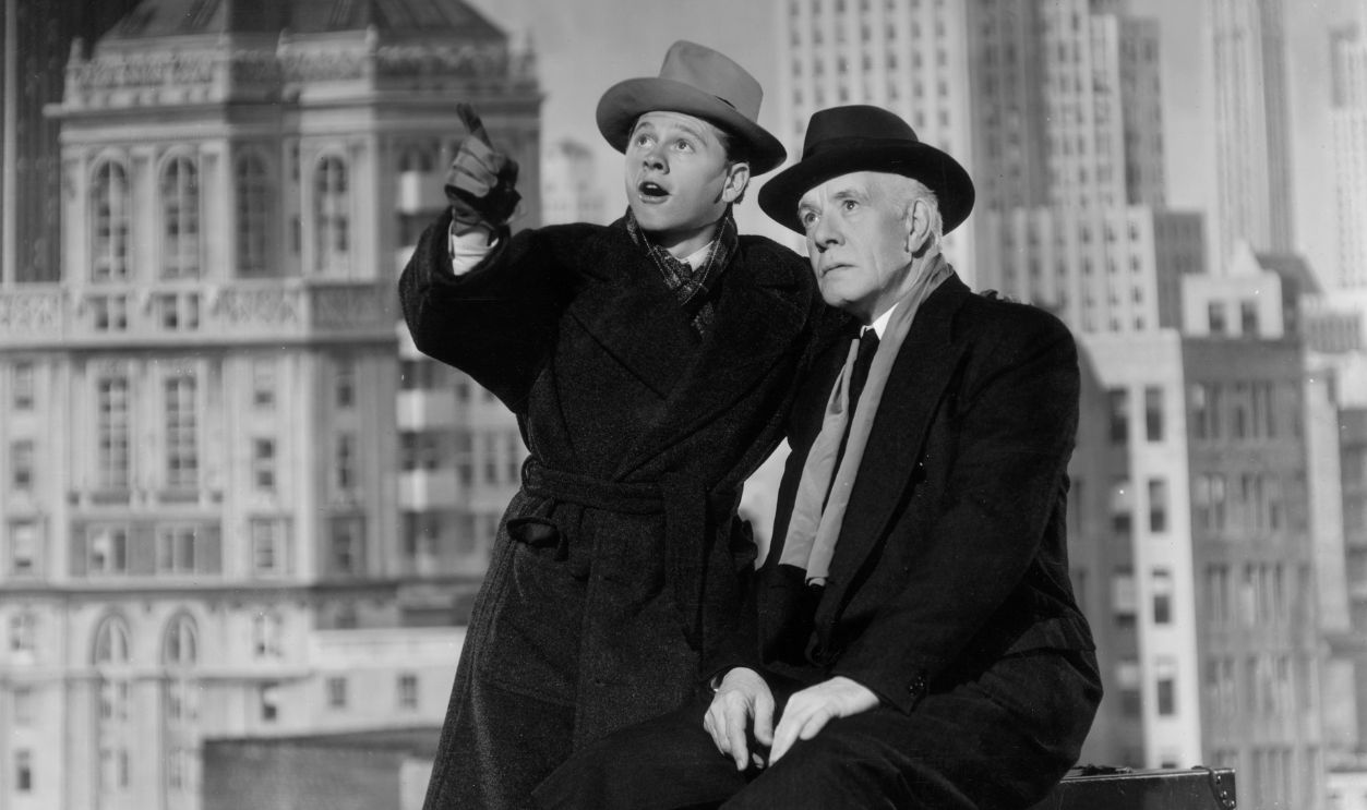 American actor Mickey Rooney (left) points upwards as American actor Lewis Stone looks on, in a still from director George B. Seitz's film, 'Andy Hardy Meets a Debutante'. The actors are surrounded by luggage in a scene against a backdrop of skyscrapers.