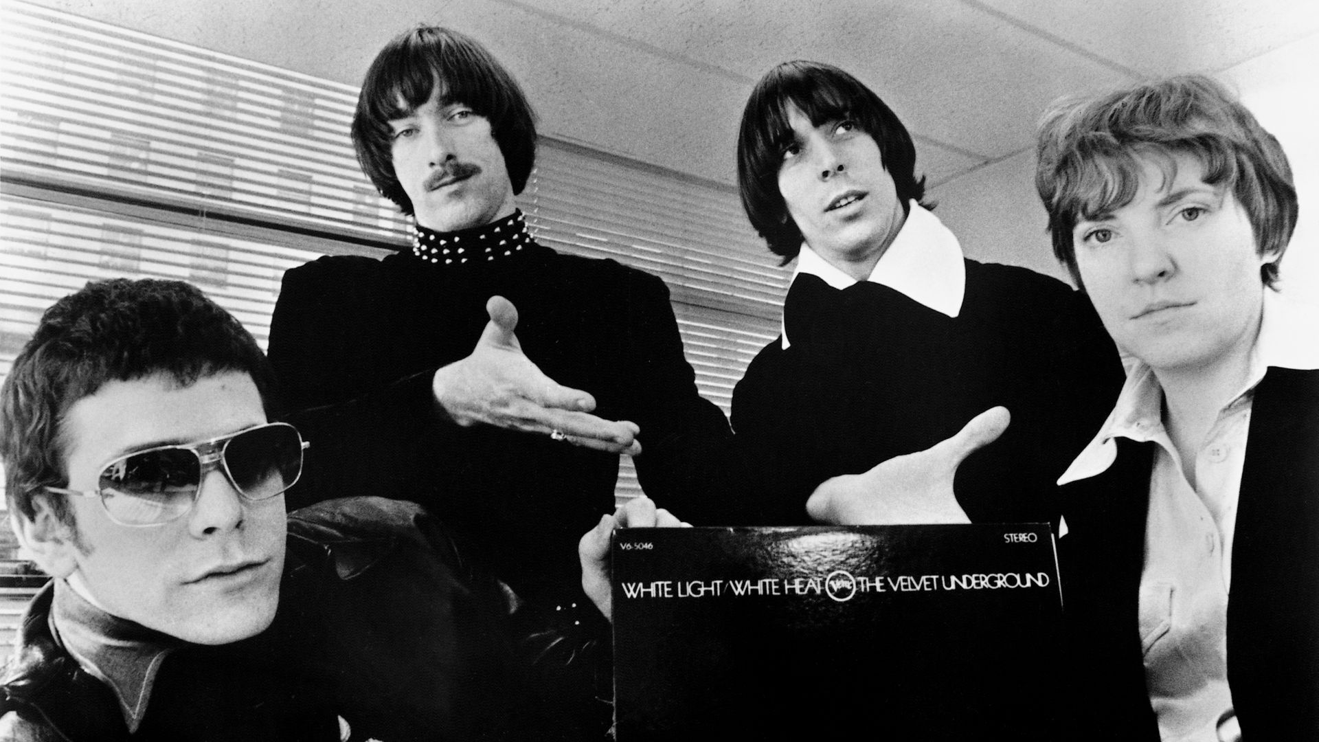 A publicity photo of the American rock band The Velvet Underground circa 1968, promoting their second album White Light/White Heat. The band members are positioned around a copy of the album, with the text of the title visible against a black background. 