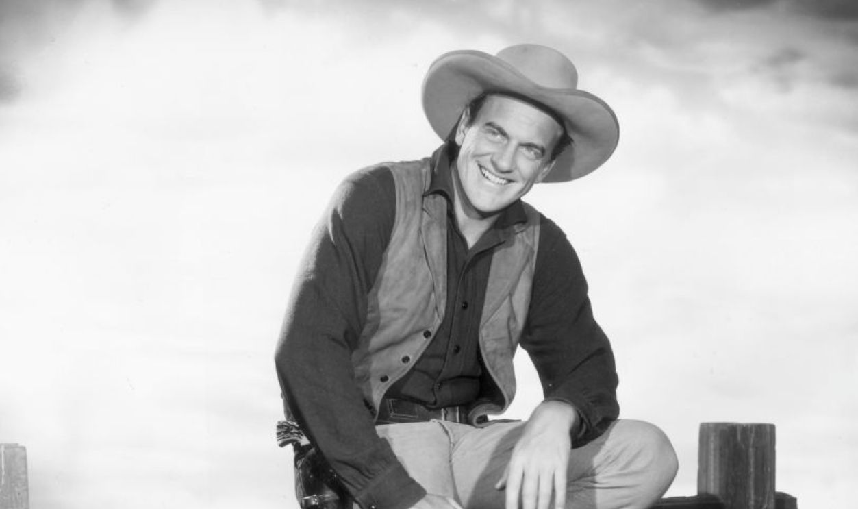 circa 1955: American actor James Arness wears a Western costume while sitting on a fence in a promotional portrait for the television series, 'Gunsmoke,' 1950s. Arness played Marshall Matt Dillon.