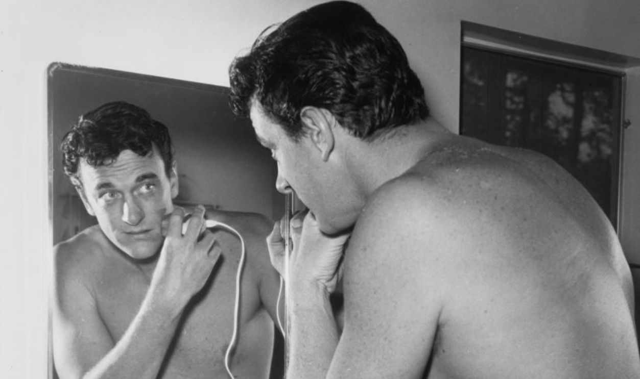 American actor James Arness (1923 – 2011) shaving in front of a mirror with an electric device, US, circa 1960.