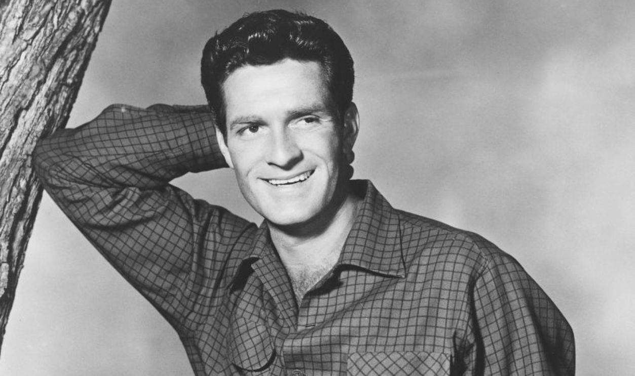 Hugh O'Brian, US actor, wearing grey trousers and a dark fine check shirt, leaning with his right elbow against a tree trunk, his left hand in the pocket of his trousers, in a studio portrait, against a light background, circa 1955.