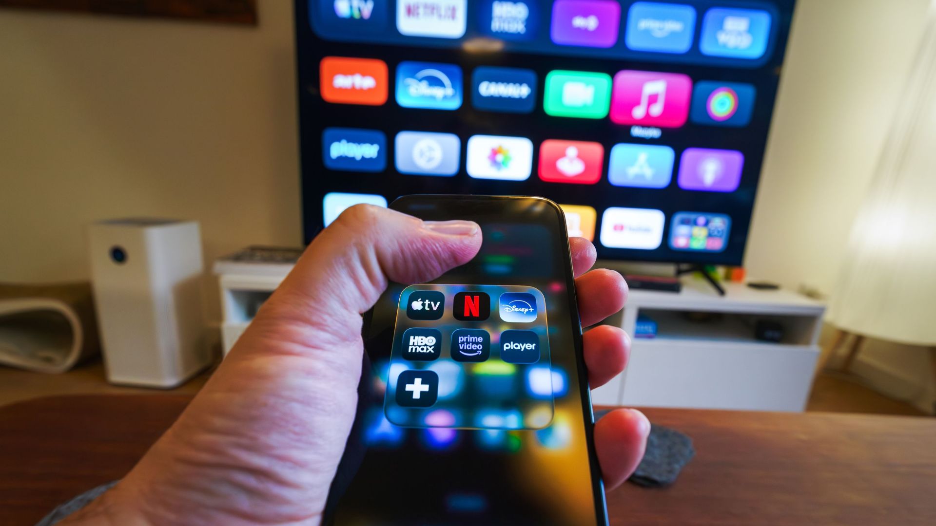 Close-up of a hand holding a phone displaying streaming apps in front of a TV with multiple app icons.