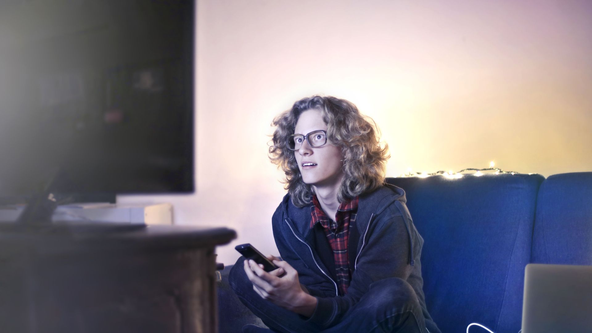 Concentrated male with long hair sitting on comfortable sofa at home and messaging on social media via cellphone while watching movie on TV with opened mouth