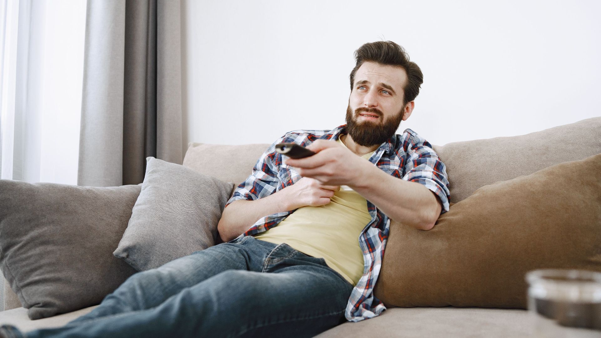 Casual bearded man lounging on a sofa, watching TV and relaxing in a cozy living room.