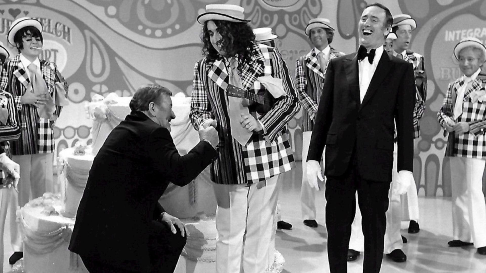 Publicity photo from the 100th episode of the television program Rowan & Martin's Laugh-In.  Guest stars are John Wayne and Tiny Tim. Host Dick Martin is pictured, along with past and present cast members from the show.  Visible in the background are Ruth
