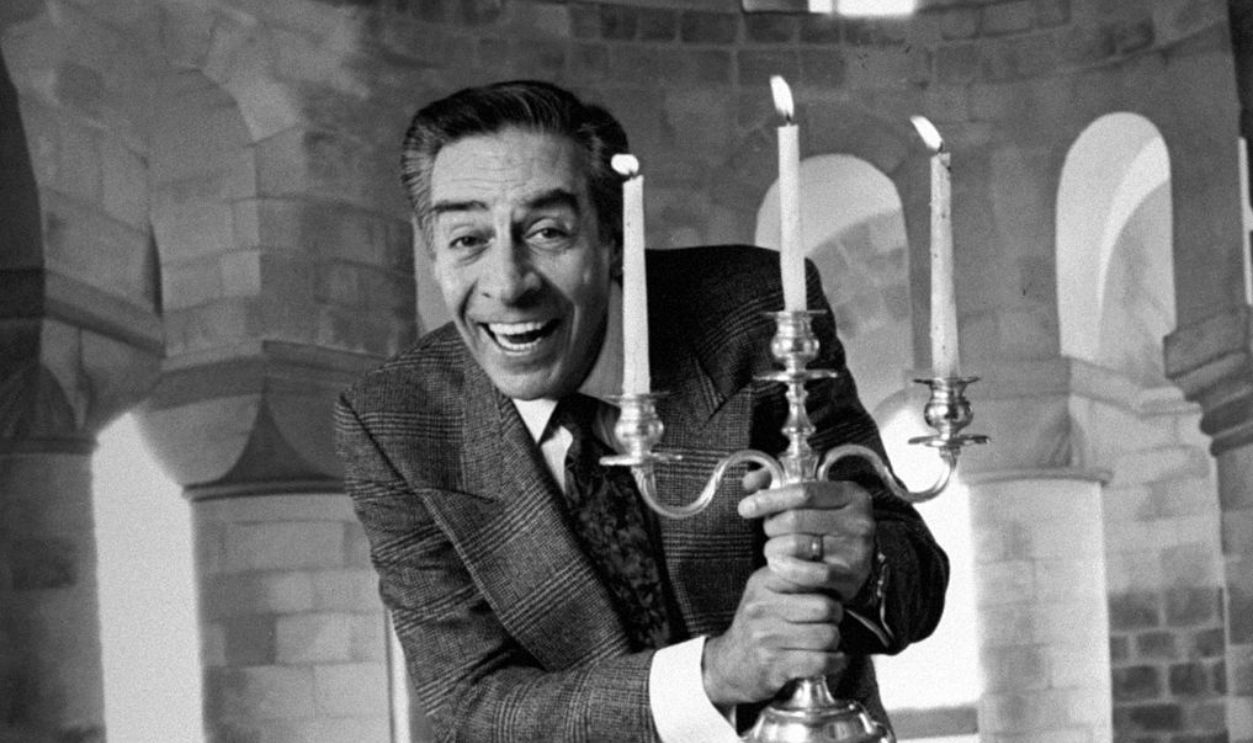 Actor Jerry Orbach holding a silver candelabra w. 3 lit candles in front of backdrop at studio, acting like the character Lumiere, the candlestick, for which he did the voice in the animated film Beauty and the Beast, October 1991.