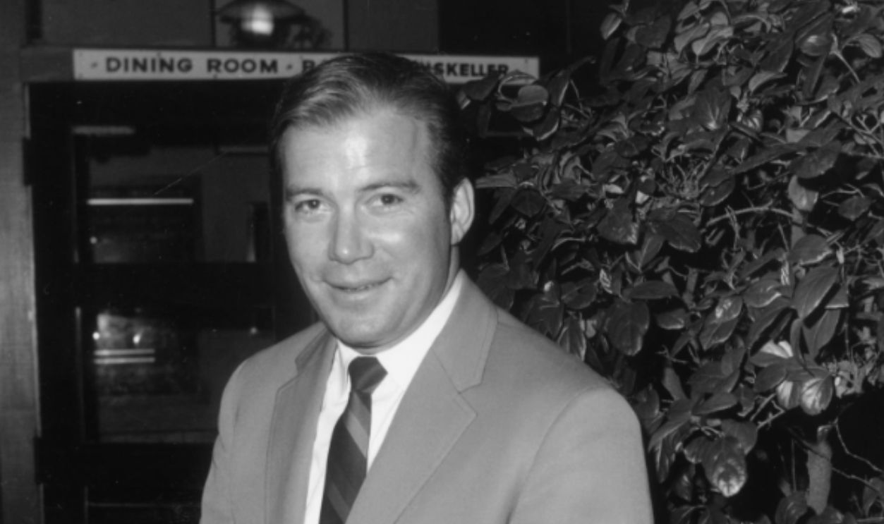 1968: Canadian-born actor William Shatner, wearing a light-colored suit and striped tie, clasps his hands and smiles while standing in front of a shrub indoors.