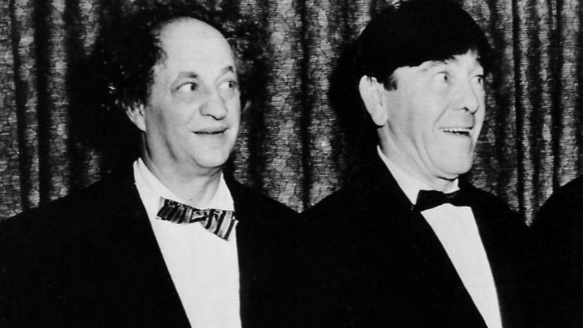 Postcard photo of The w:Three Stooges.  From left: w:Larry Fine, w:Moe Howard, and w:Joe DeRita.  w:Curly Howard, one of the original Stooges, suffered a debilitating stroke while filming in 1946 and died in 1952.  After Curly was unable to work, w:Shemp 
