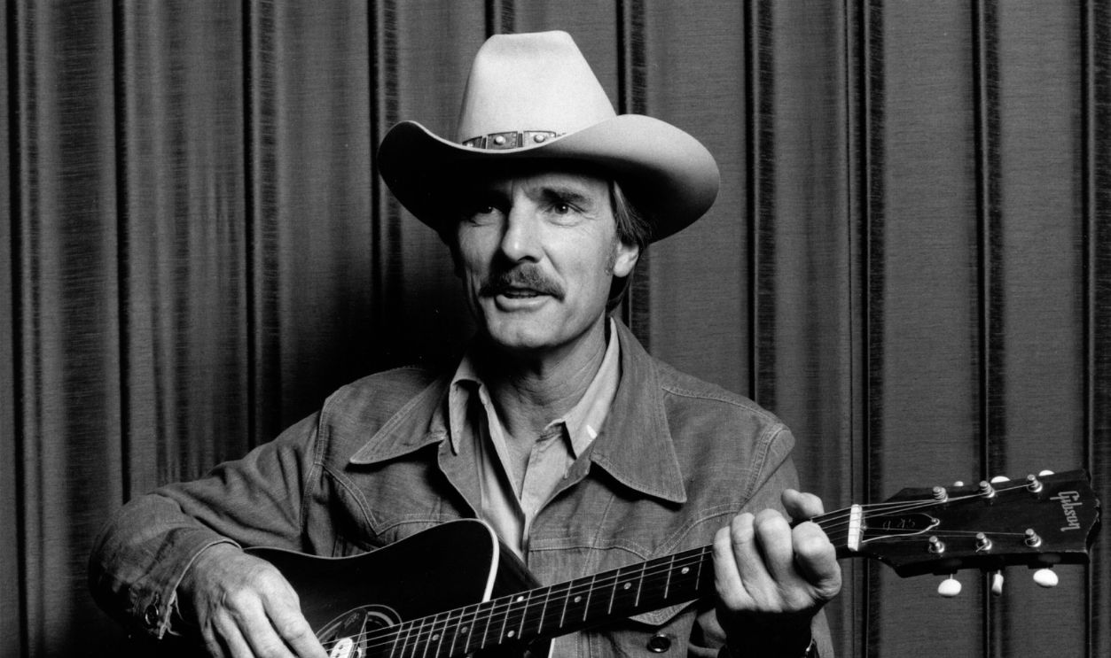 American actor Dennis Weaver wearing a cowboy hat and playing the guitar