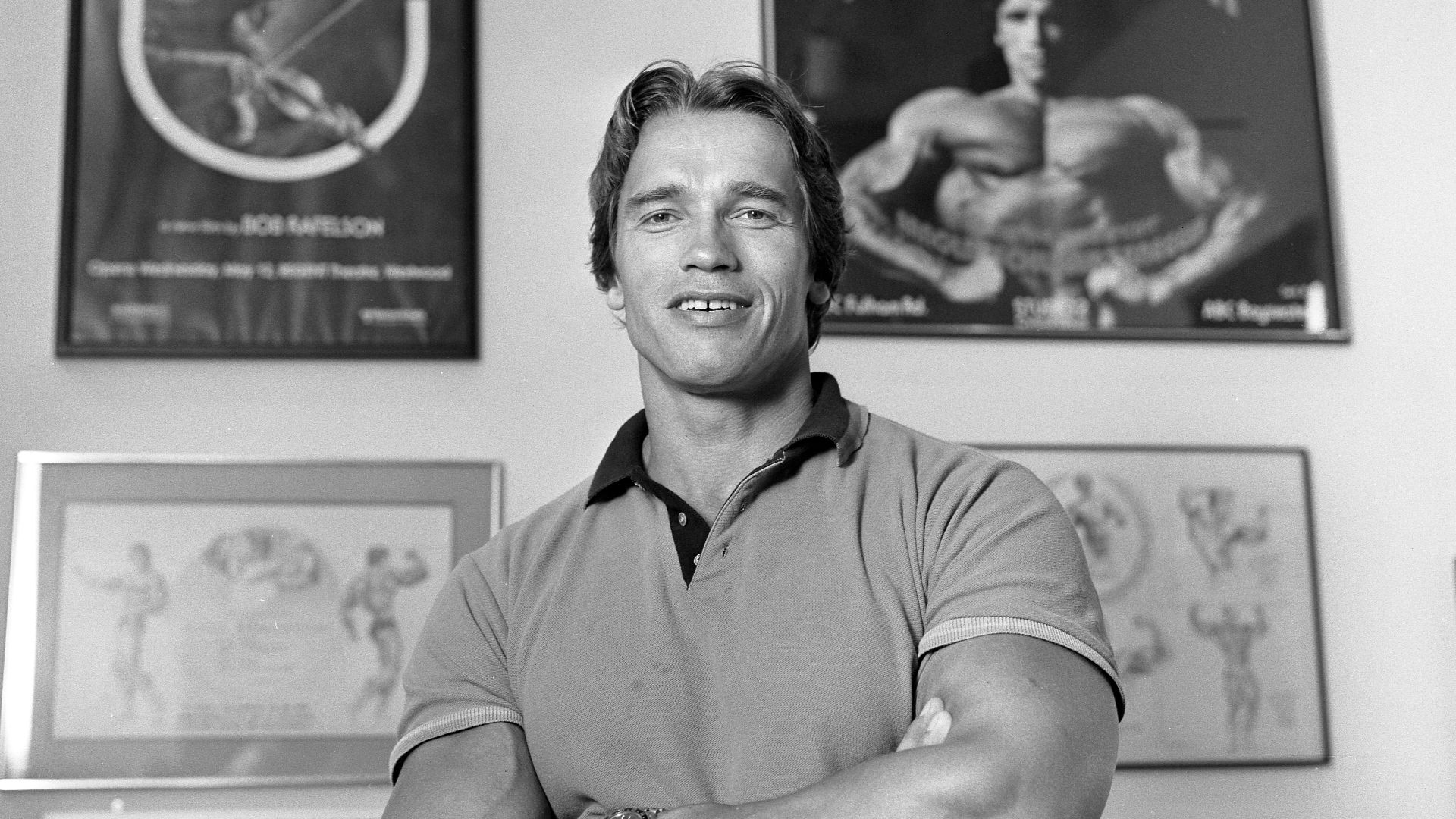 Arnold Schwarzenegger standing with arms crossed in front of some photos of him and posters for movies that he appeared in