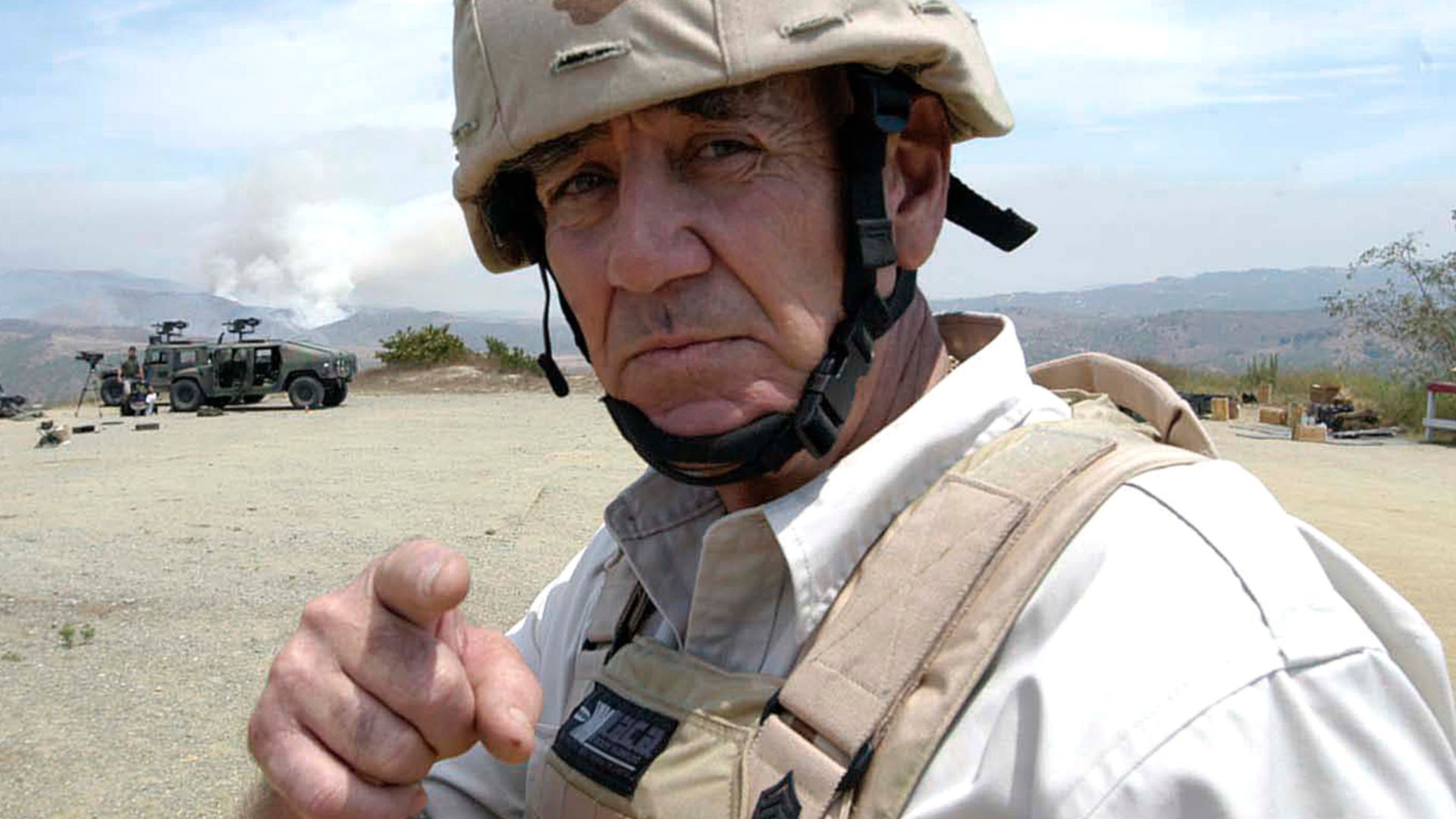 Retired Gunnery Sgt. R. Lee Ermey, host of the History Channel's �Mail Call,� poses for the camera while members of the advanced infantry training battalion fire from a vehicle-mounted, MK-19 grenade launcher during their first week of the course.