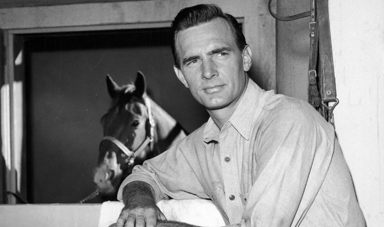 Portrait of American actor Dennis Weaver, propping his foot on top of a bench and leaning on his knee, posing in a barn in front of a horse in a stall. Weaver was the star of various western films and the television series, 'Gunsmoke.'