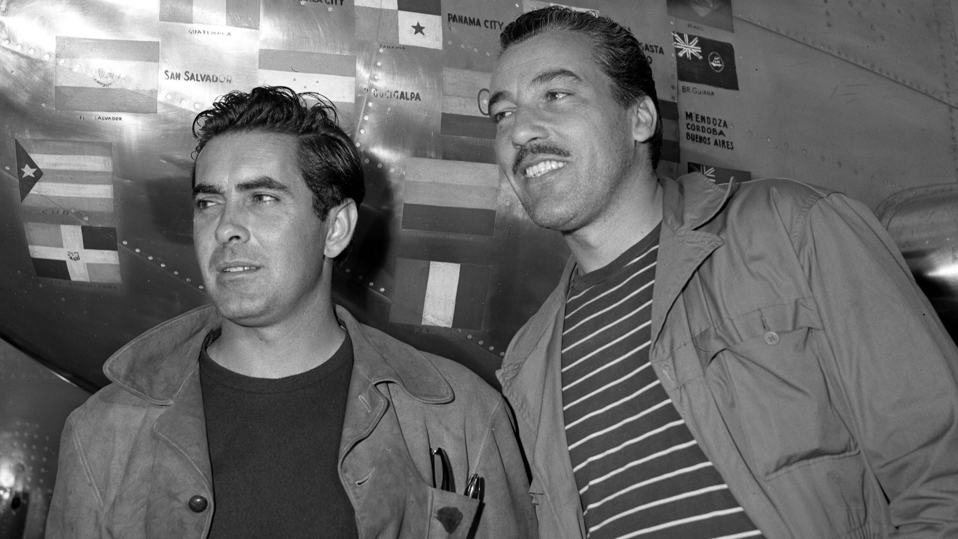 Tyrone Power, left, and Cesar Romero pictured beside Power's plane on return from Latin-American good-will tour. Flags of various nations visited by Power are painted on side of plane.
