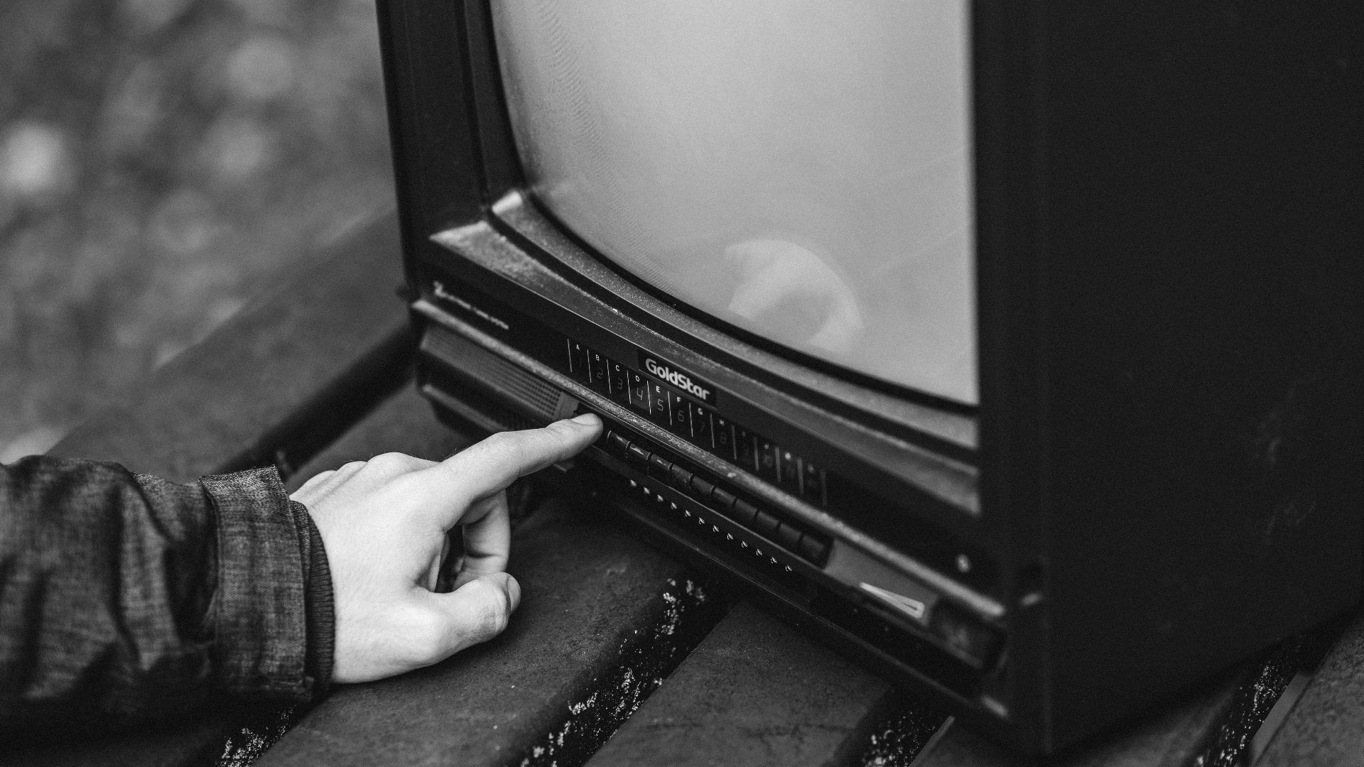 Black and white of unrecognizable male pressing buttons on old fashioned television placed on wooden table in nature on blurred background