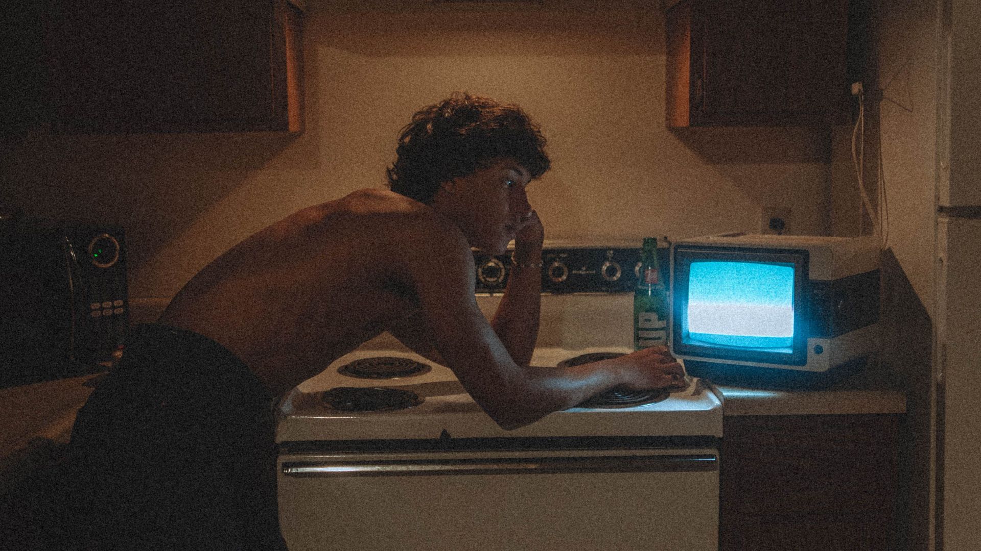 Shirtless man leaning thoughtfully over stove with a vintage TV in a dimly lit kitchen.