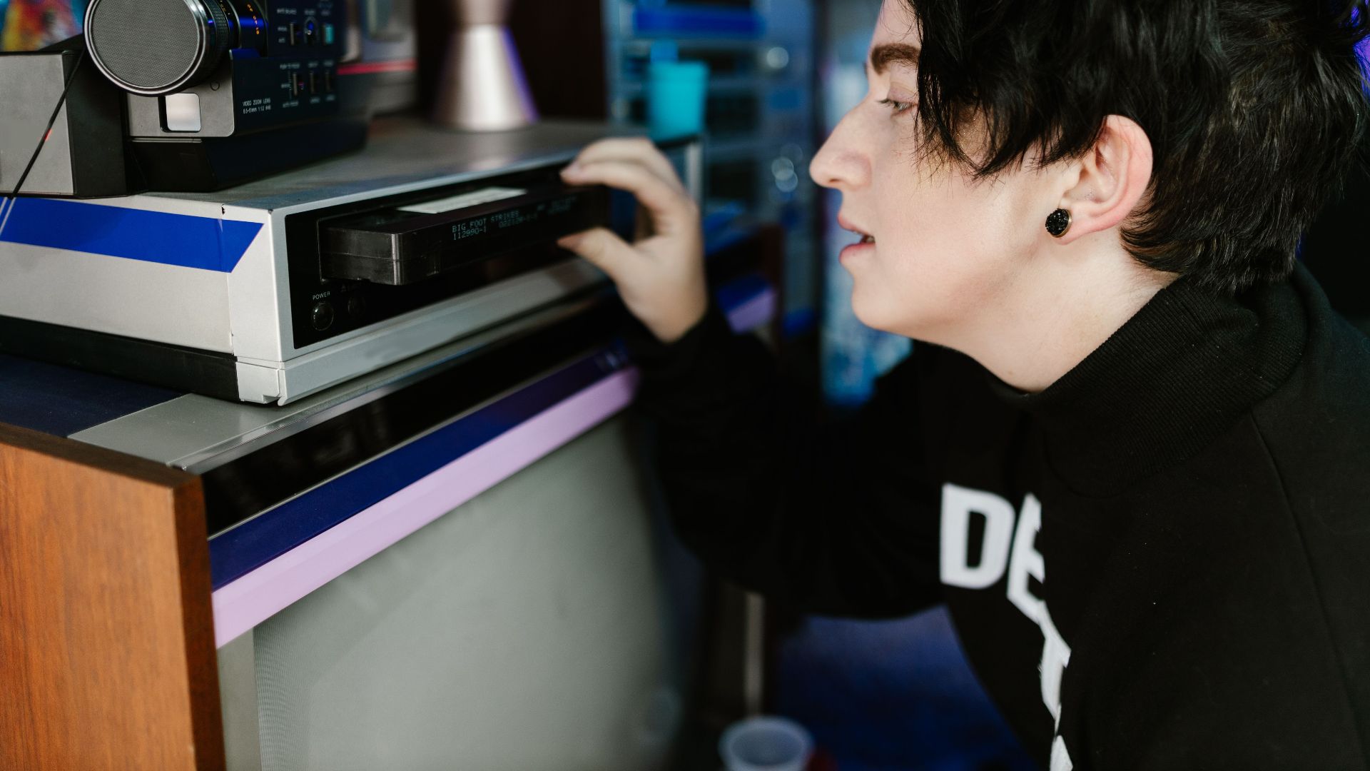 A person using a vintage VHS player, surrounded by retro decor and neon lighting.