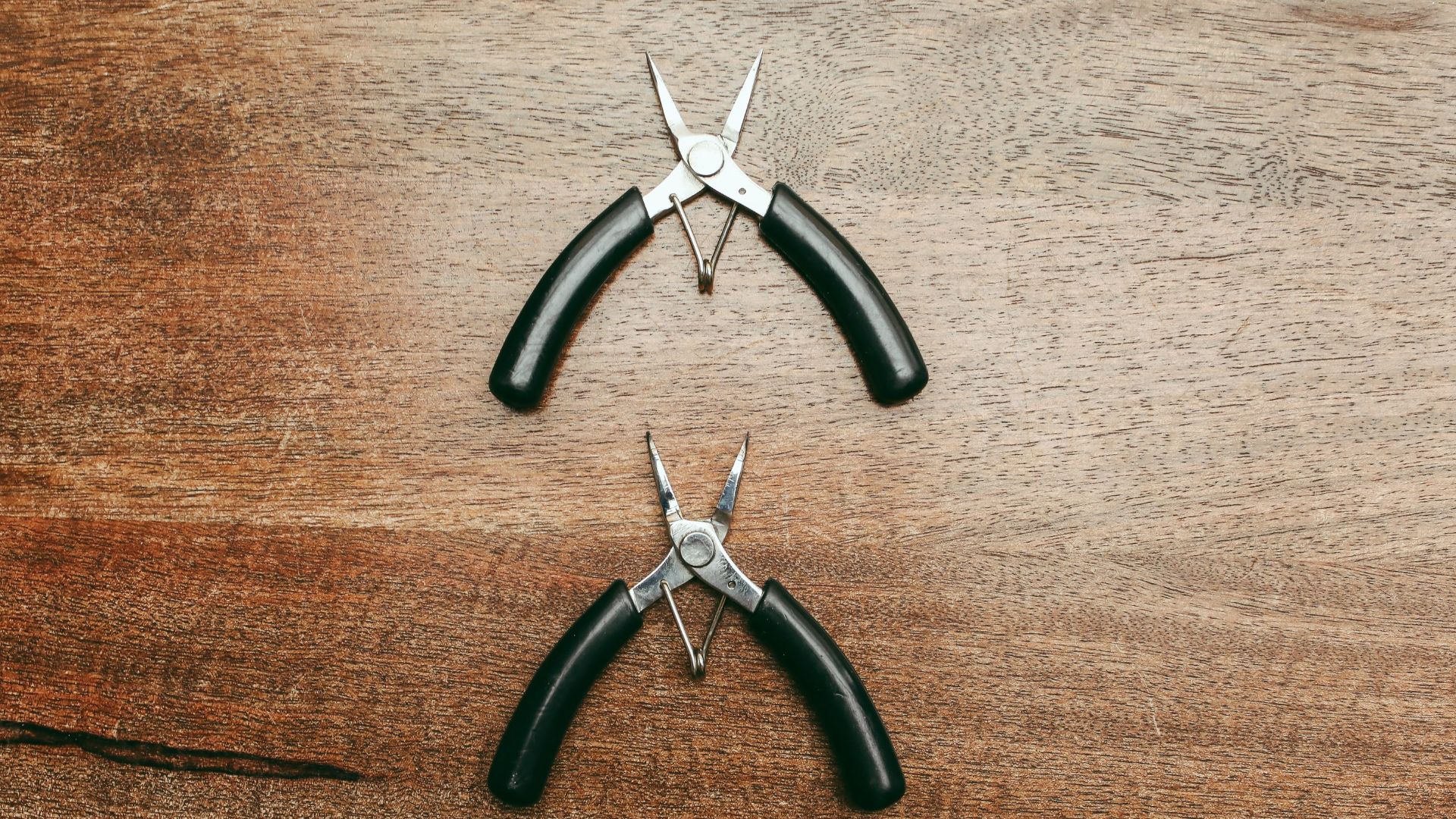 Close-up of two needle-nose pliers on a wooden tabletop, showcasing stainless steel construction and black handles.