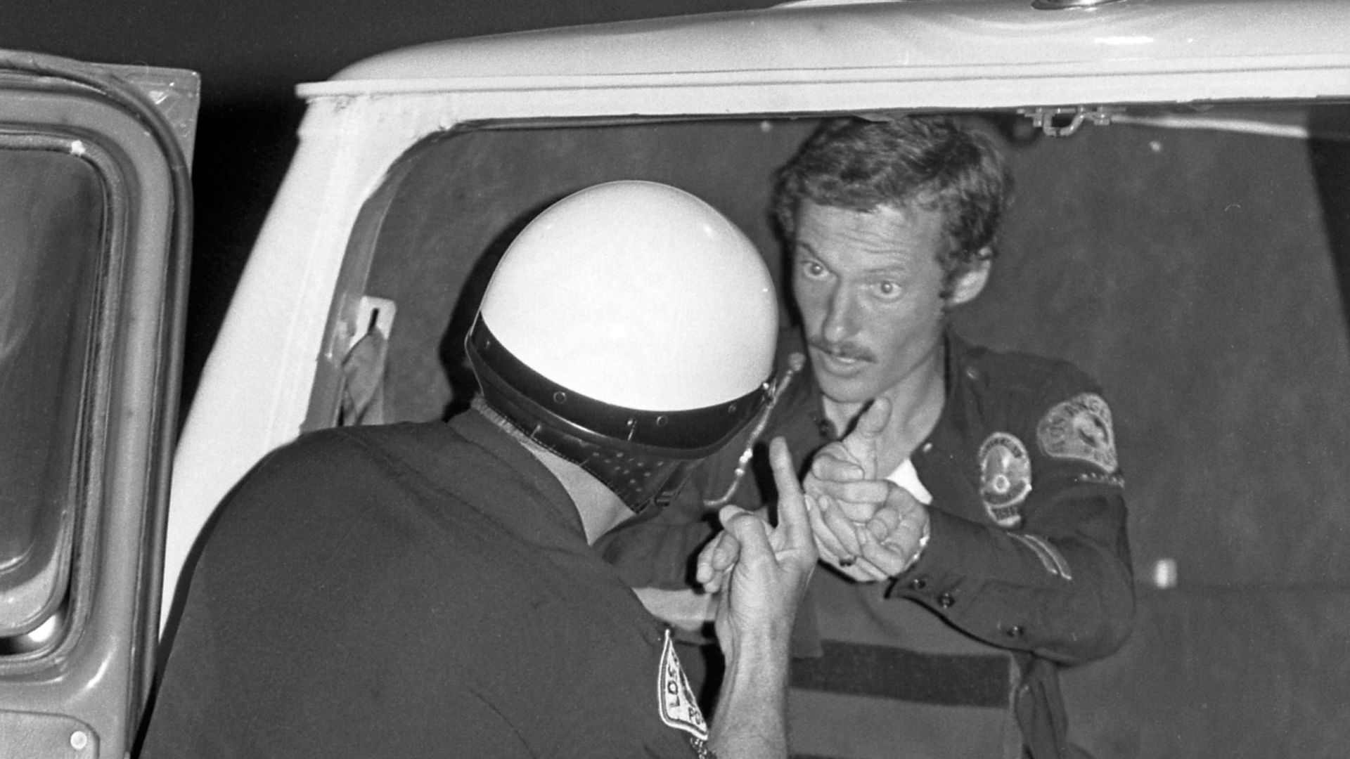 REAL-LIFE DRAMA—Officer Kenneth Osmond (facing camera), who for years played a part on the Leave It to Beaver television series, tells a fellow officer how he escaped death when a fleeing car theft suspect shot him three times. He was saved by bulletproof