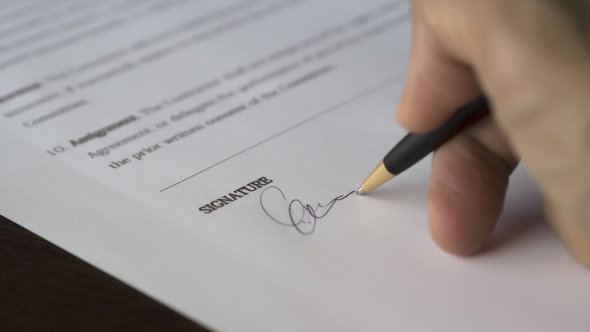 A hand signs a formal contract with a pen on a wooden desk.