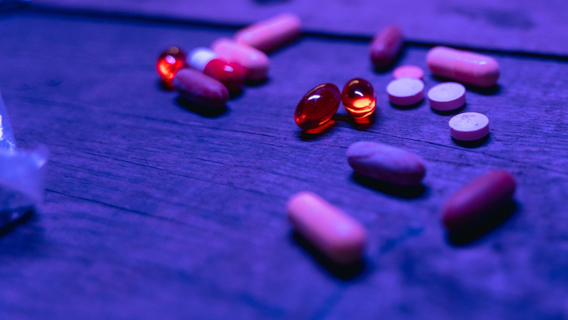 A variety of capsules and pills scattered on a wooden surface under blue lighting.