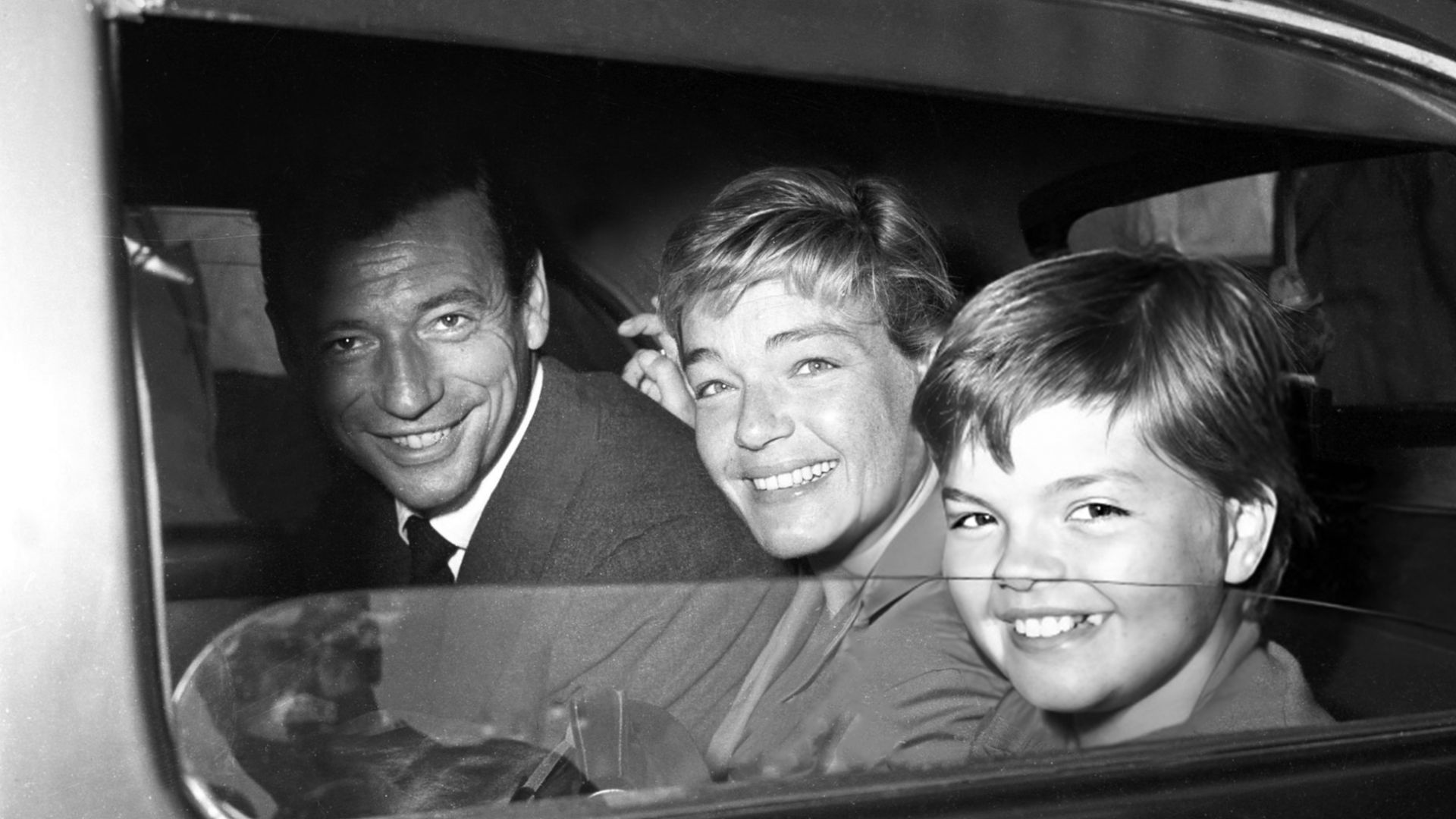 À l'intérieur et à l'arrière d'une automobile sombre dont la vitre est largement descendue, Yves Montand, Simone Signoret et au premier plan, Catherine Allégret sourient largement, la tête et le regard tournés vers l'objectif. Ils portent des vêtements gr