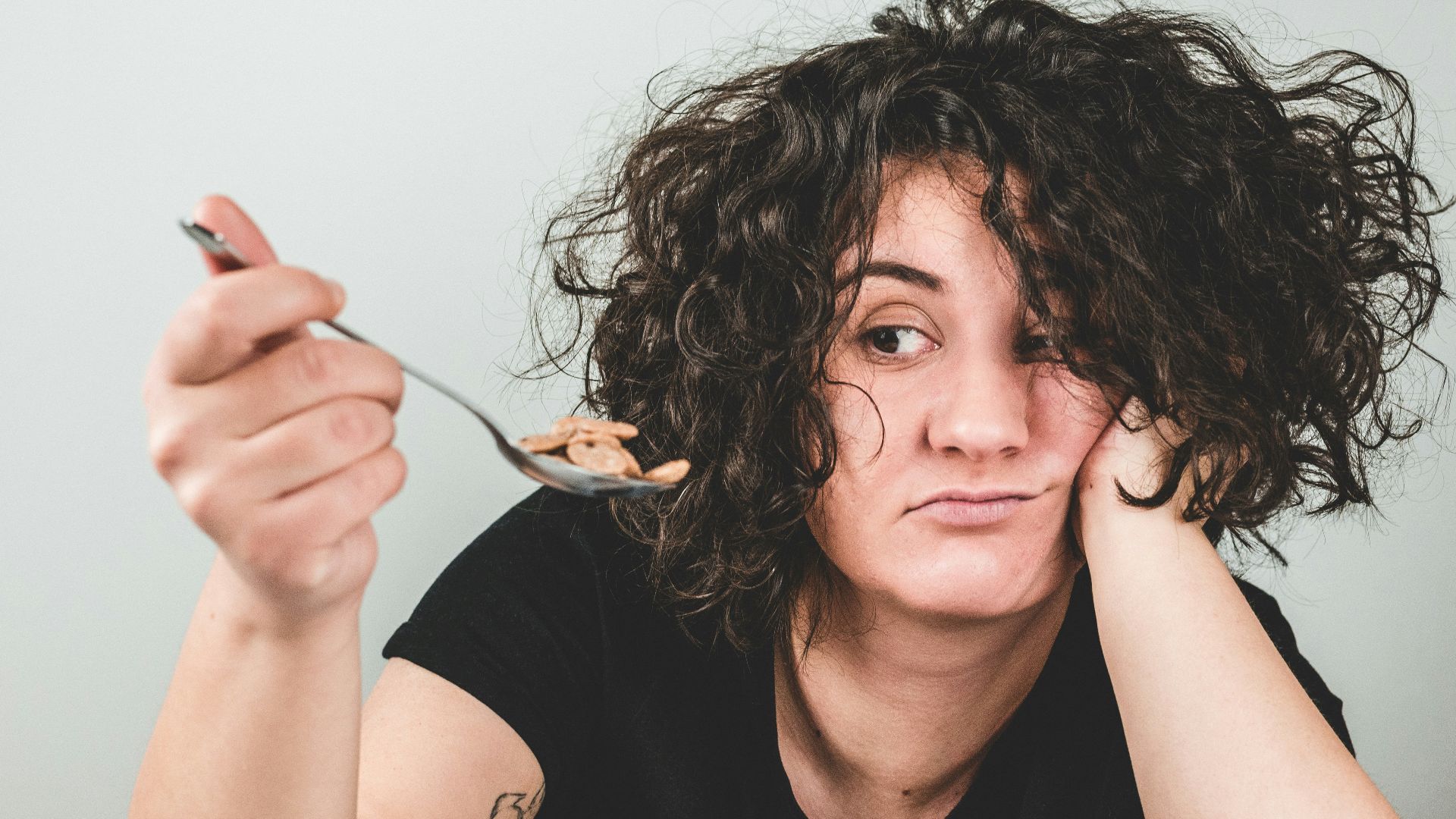 woman with messy hair wearing black crew-neck t-shirt holding spoon with cereals on top