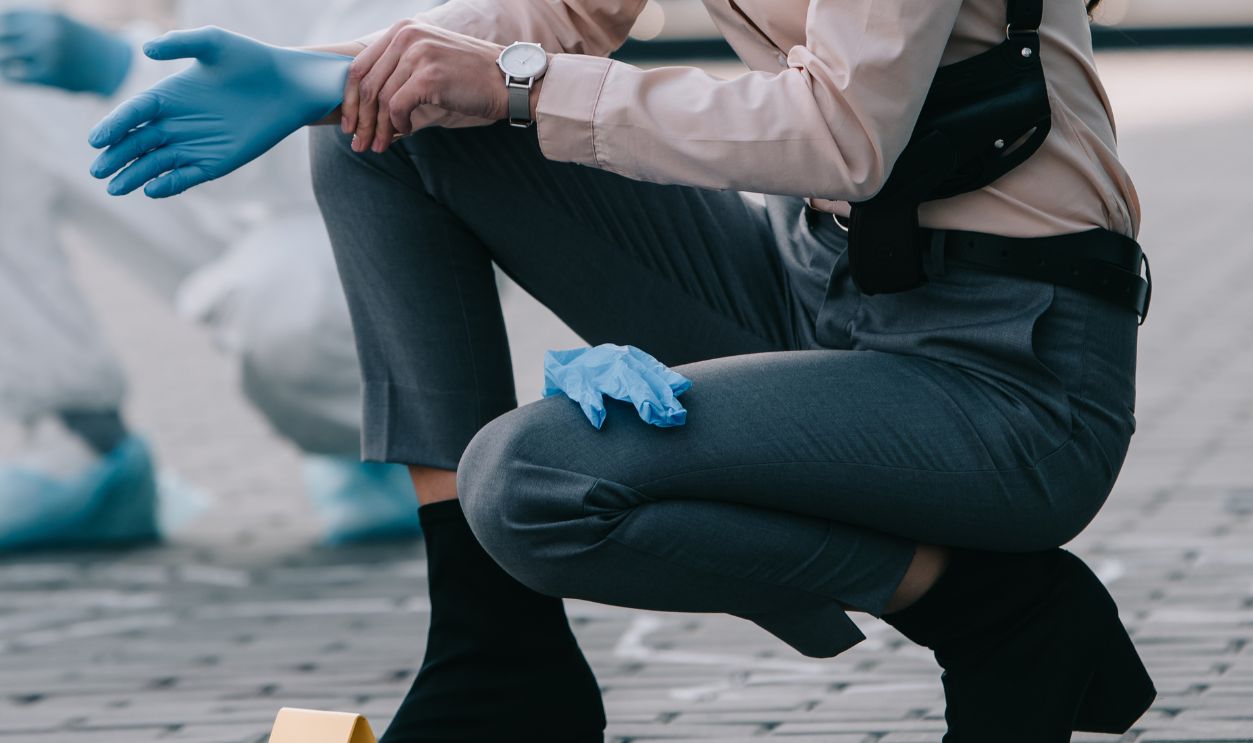 female detective in sunglasses wearing latex gloves with criminologist behind at crime scene