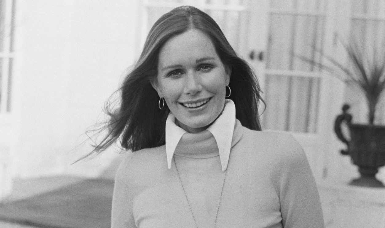 American actress Sally Kellerman wearing a turtleneck sweater with the collar of her blouse protruding over the top, with a locket hanging from a chain around her neck, posing with her hands in her pockets, United Kingdom, March 1973