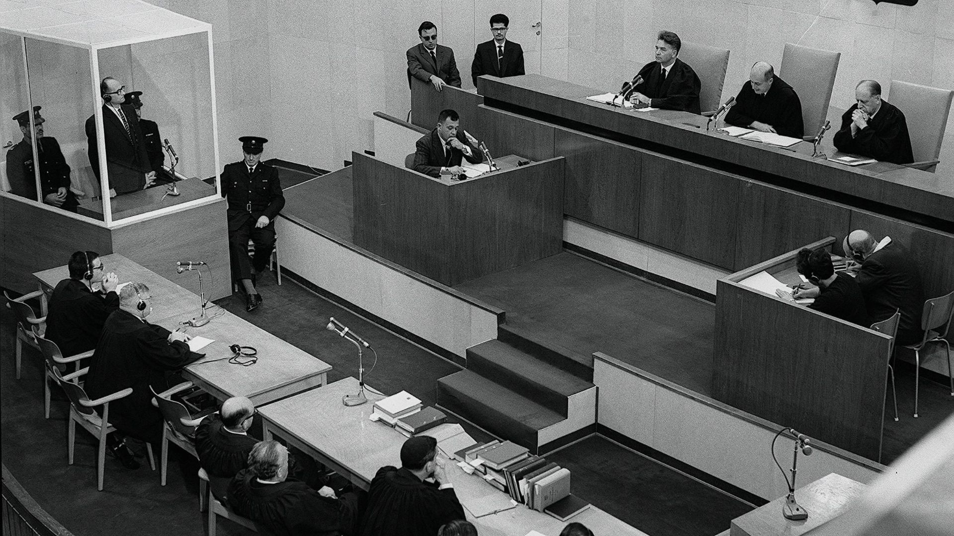 Defendant Adolf Eichmann (inside glass booth) is sentenced to death by the court at the conclusion of the Eichmann Trial. At the left table seated with two persons, the person on the right (with white hair and headphones) is defense counsel Robert Servati