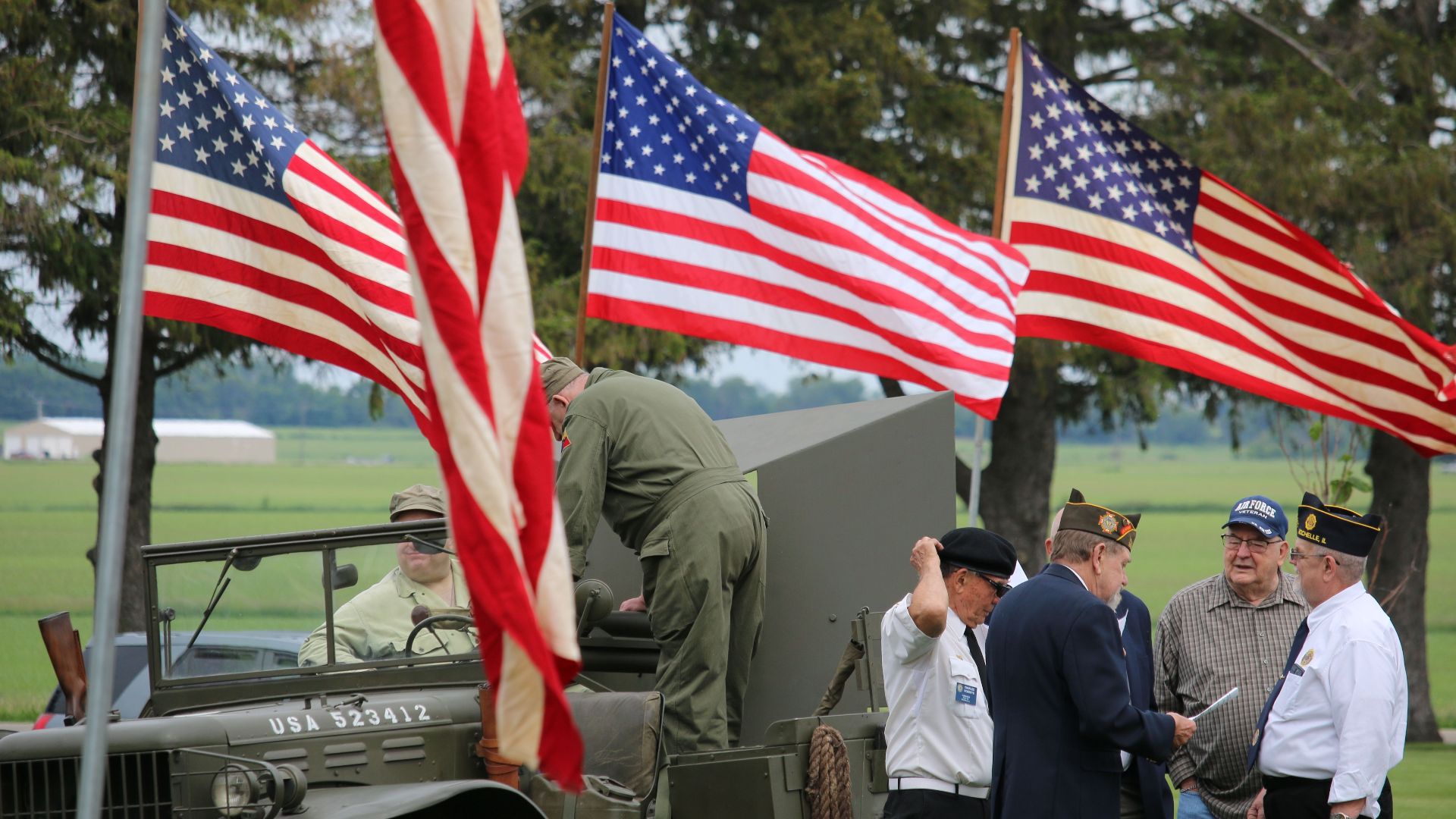 Group of veterans and soldiers by military vehicle with American flags in the background, symbolizing patriotism.