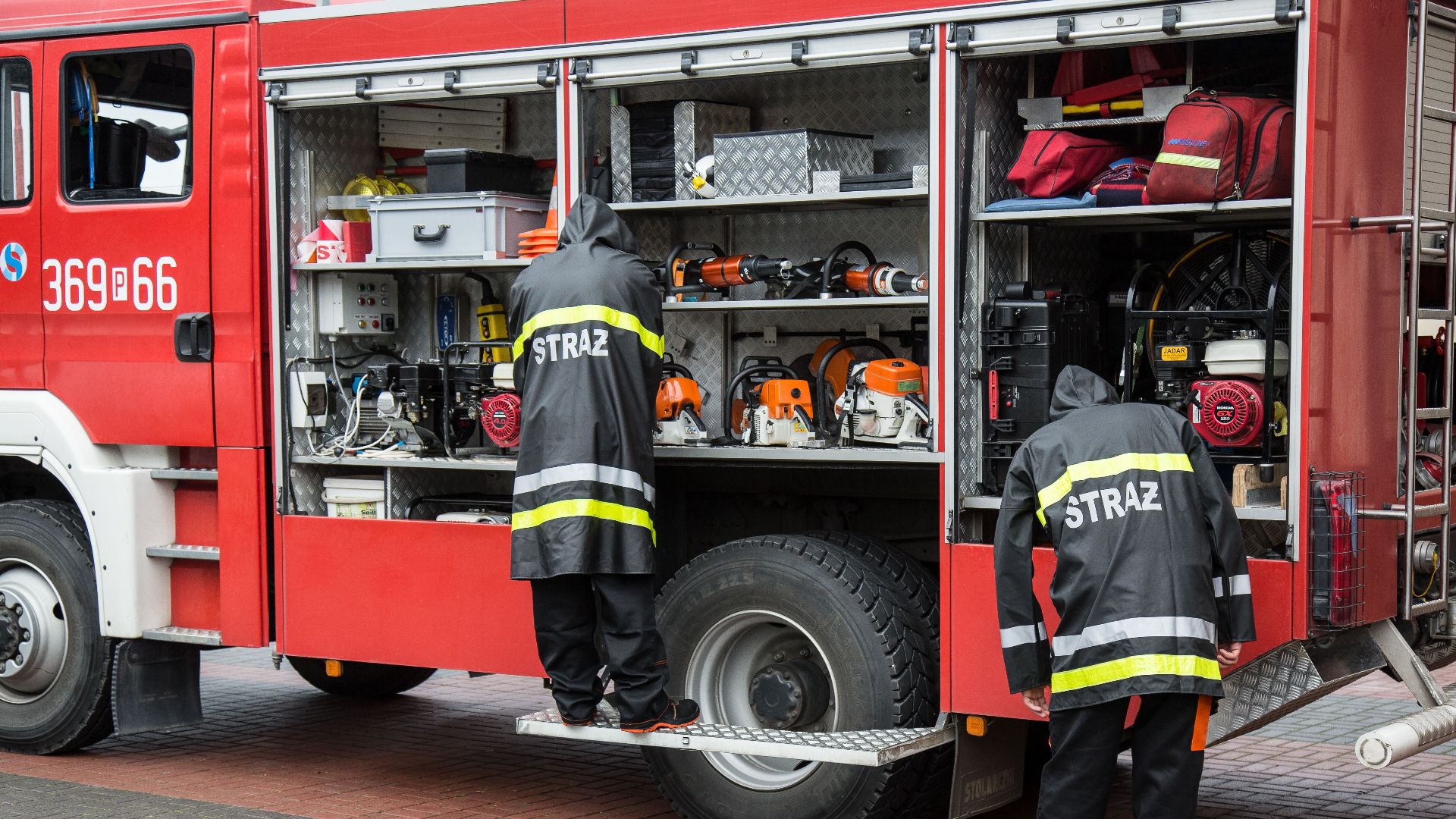Firefighters in uniform organizing equipment on a firetruck with open compartments in an urban setting.