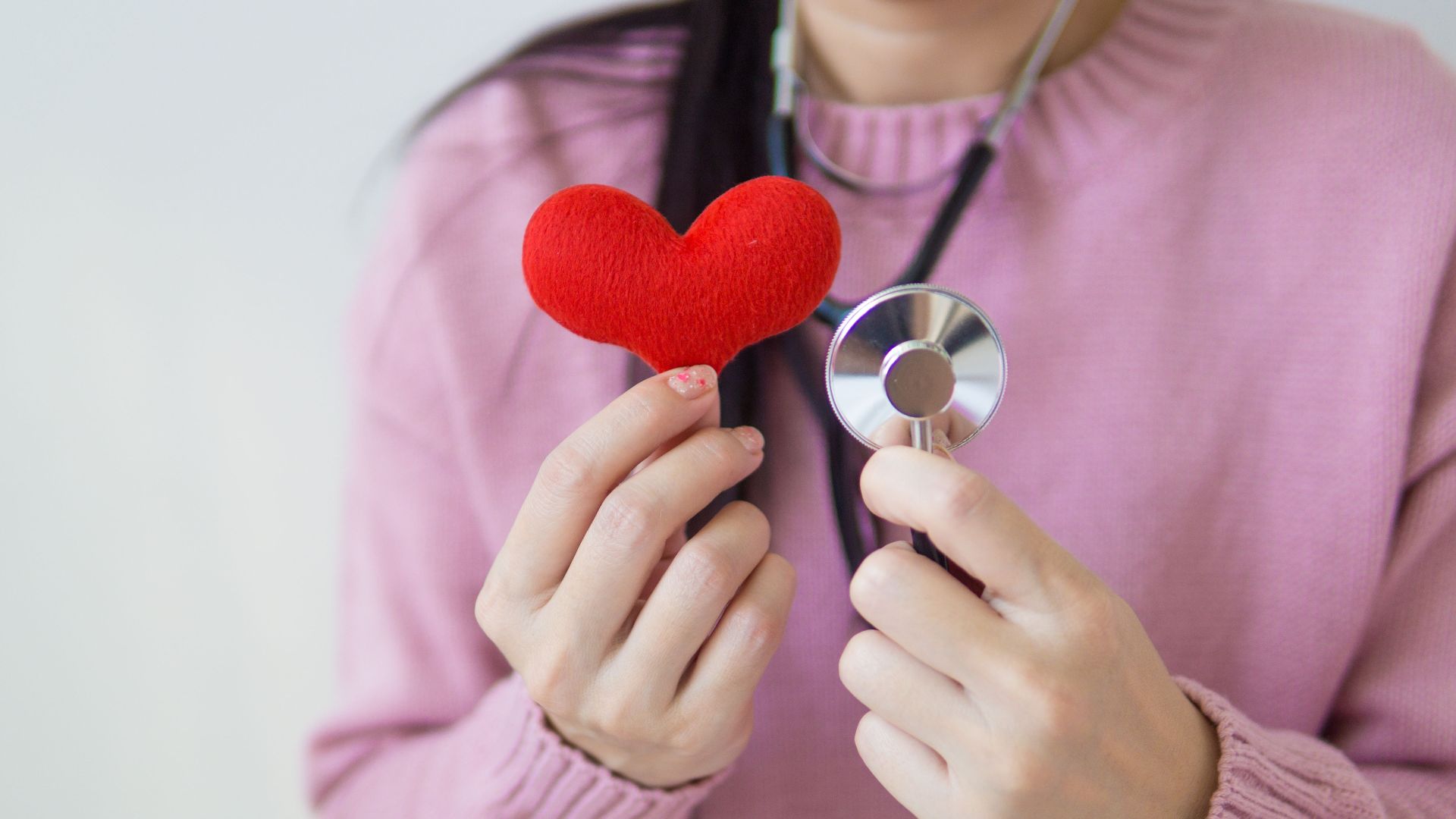 Unrecognizable female in pink sweater with stethoscope on neck standing on white background with red heart in hand in daylight