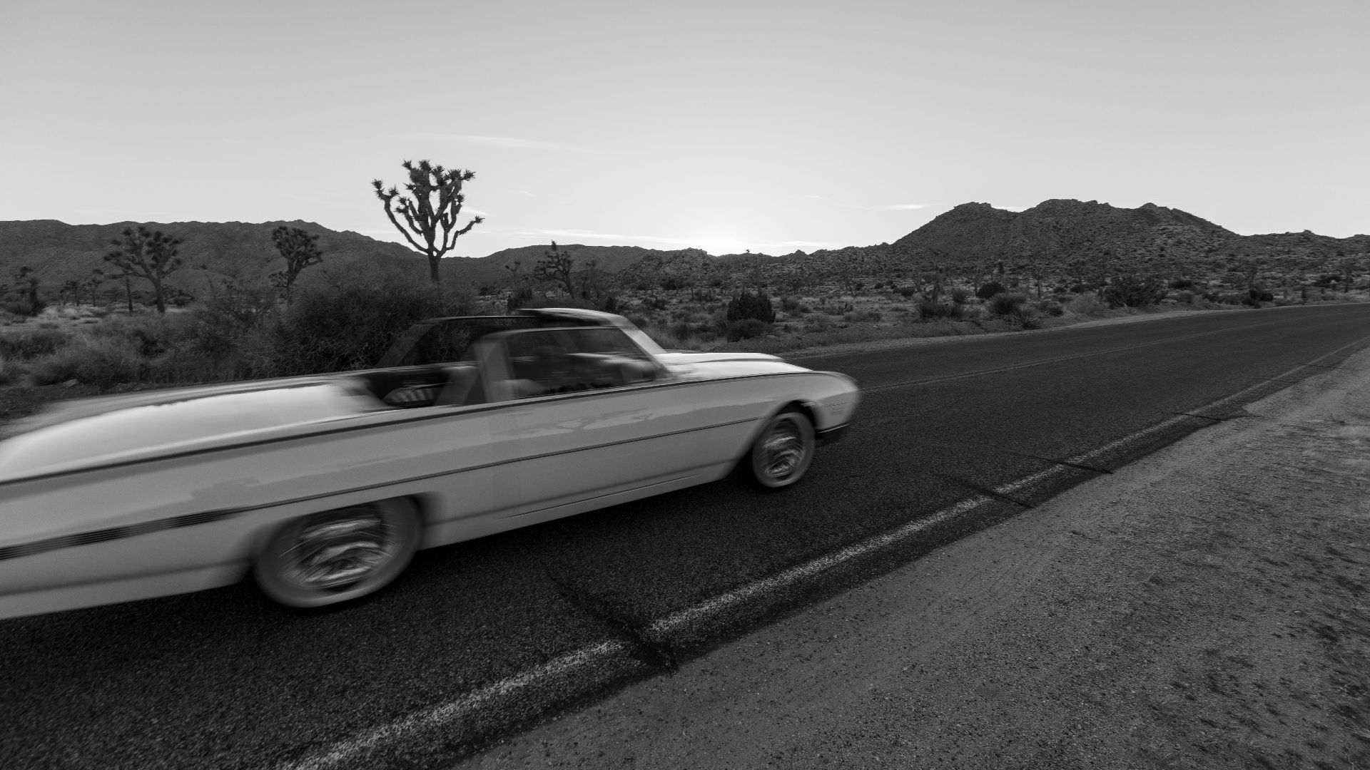 A classic convertible speeds through a desert road at twilight, showcasing motion in black and white.