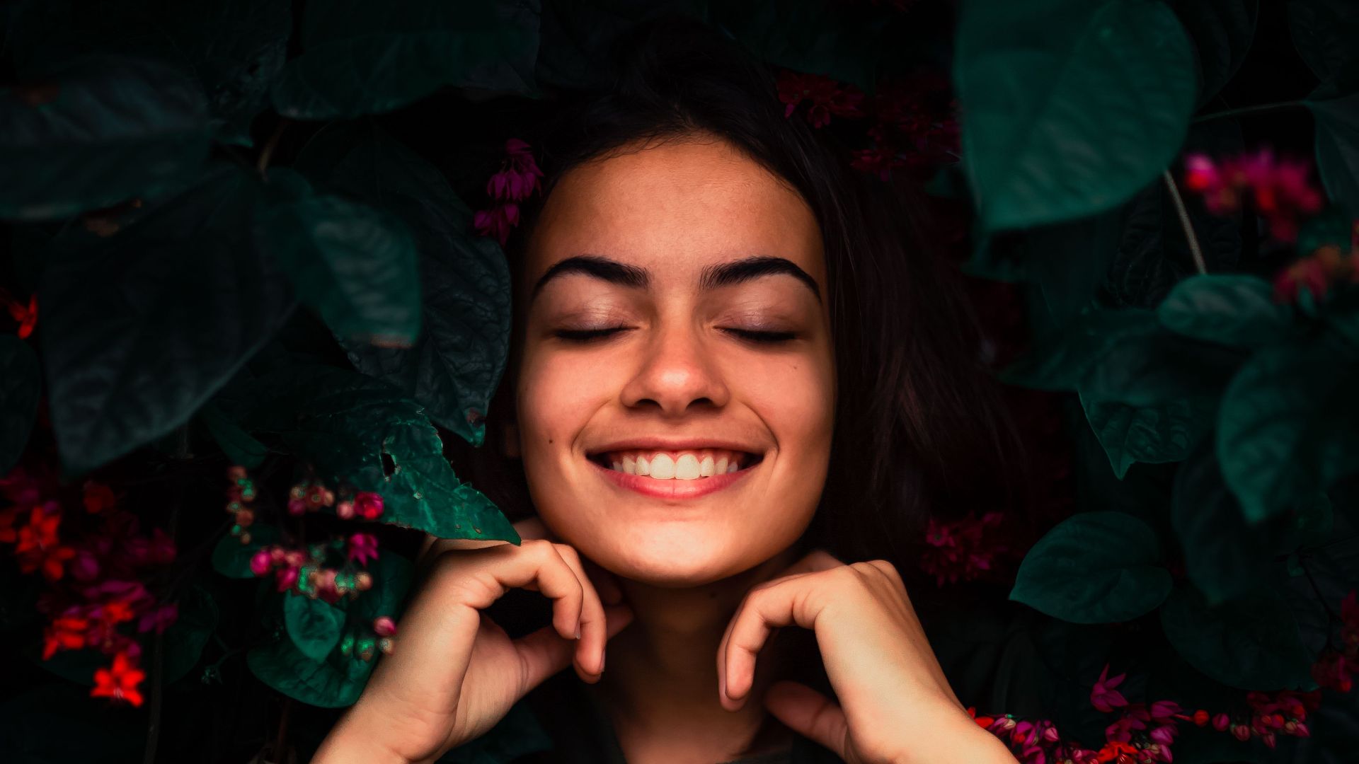 An adult woman with eyes closed smiling amidst lush greenery and vibrant flowers in an outdoor portrait.