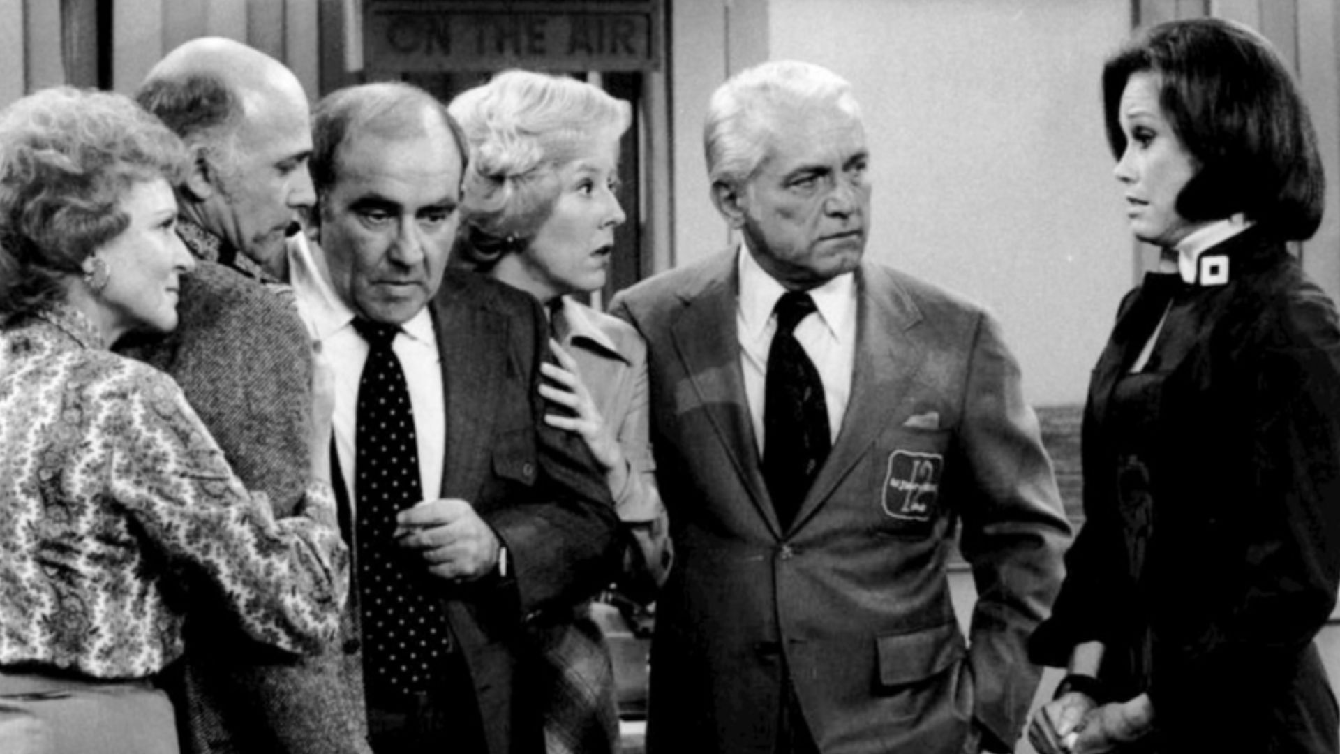 Cast photo from the television program The Mary Tyler Moore Show.  After the news that most of the WJM-TV staff has been fired, everyone gathers in the newsroom.  From left-Betty White (Sue Ann Nivens), Gavin MacLeod (Murray Slaughter), Ed Asner (Lou Gran