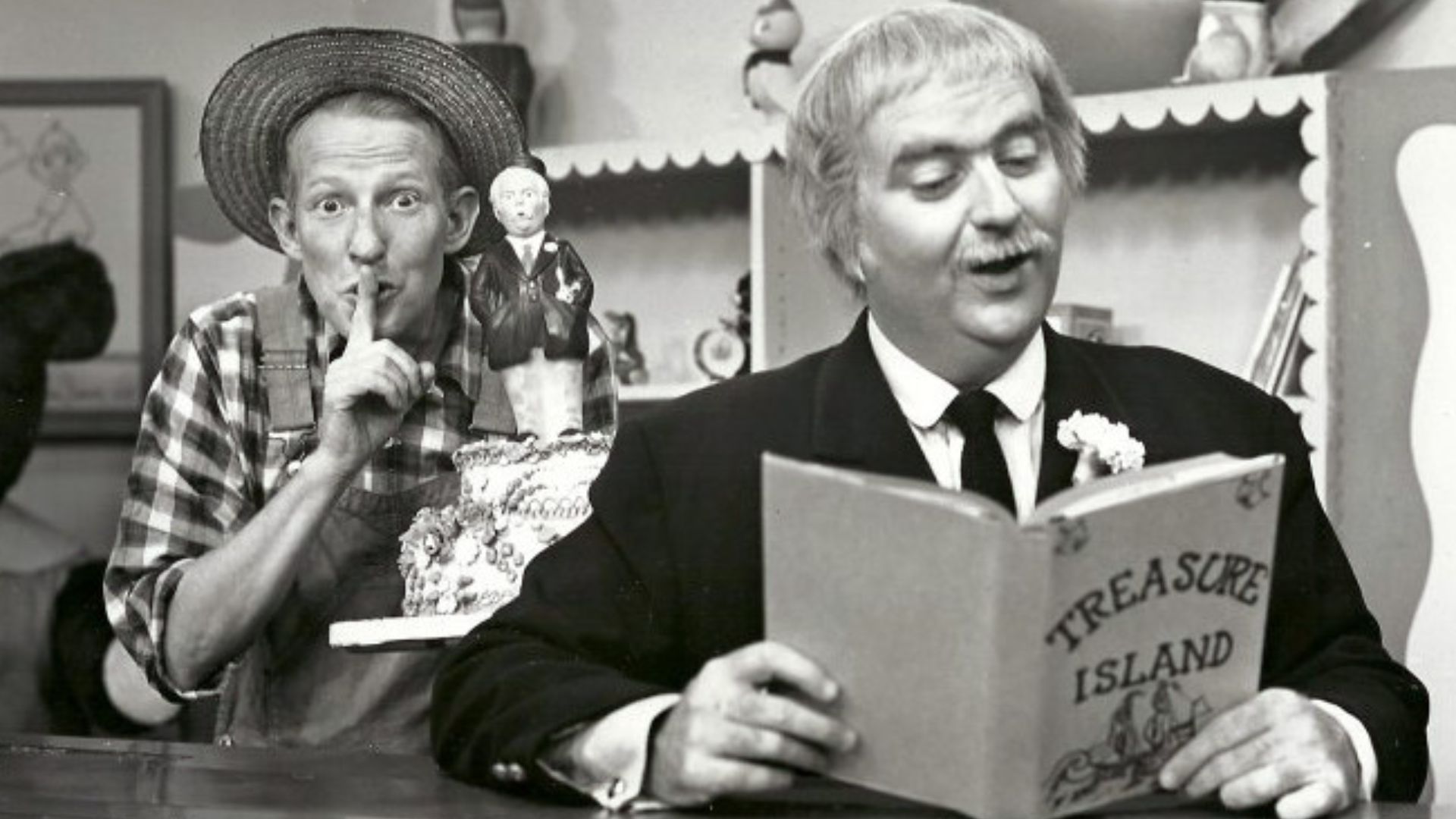 Photo of Bob Keeshan (Captain Kangaroo) reading as Hugh Brannum (Mr. Green Jeans) comes with a surprise cake to announce the fifth anniversary of the television program Captain Kangaroo.