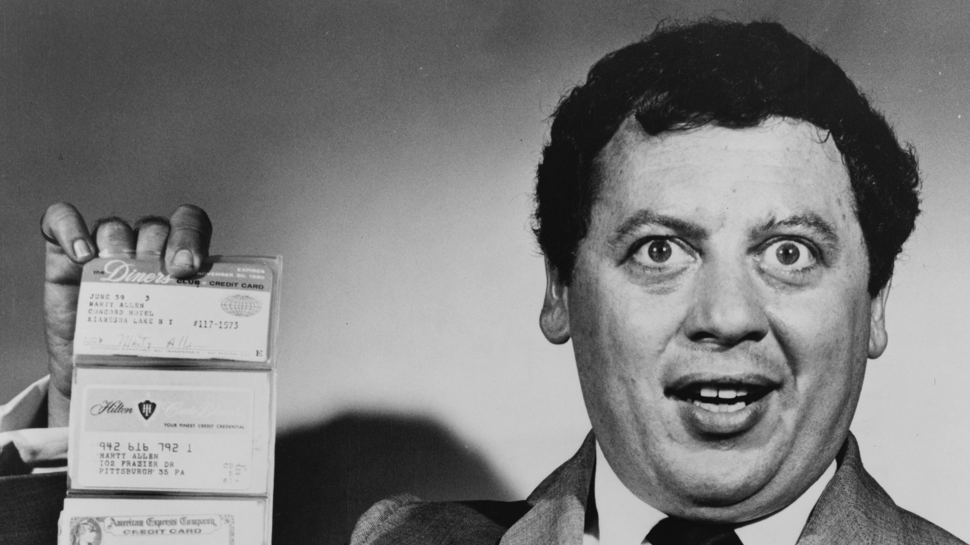 Marty Allen, American comedian, holding wallet with six cards extended in accordion fashion. Visible from top to bottom are